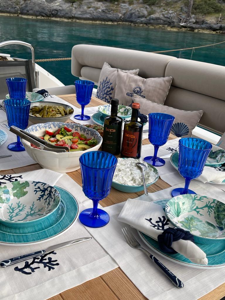 Deniz kenarında bir masa ve masa üzerinde yemek servisine hazır boş tabaklar, mavi bardaklar ve şişede zeytinyağları yer alıyor